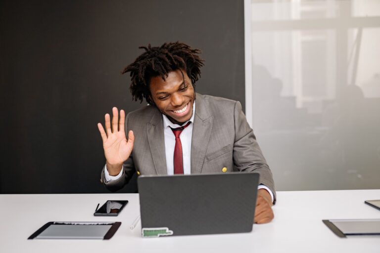 Professional making a video call, waving hand at laptop in modern office setting.