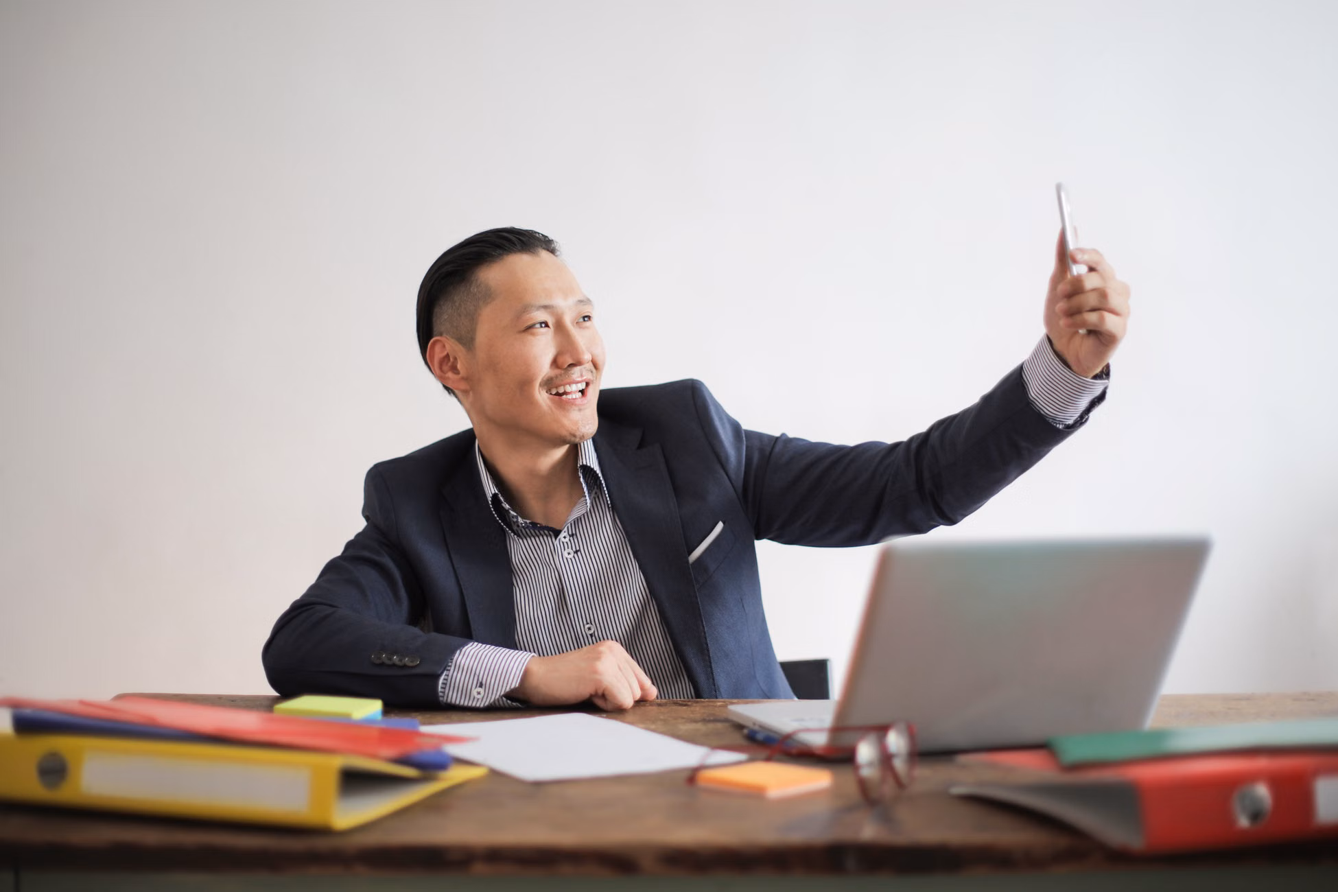 Smiling businessman taking a selfie with a smartphone at a desk with laptop and folders.
