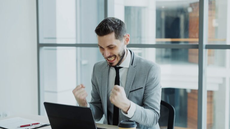Man in grey suit celebrating success at his desk with a laptop.